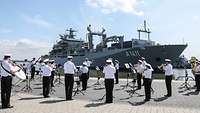 Soldaten in dunkelblauer Marineuniform musizieren an einer Pier vor einem großen gauen Kriegsschiff.