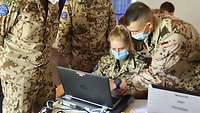 Eine Soldatin sitzt vor einem Laptop. Mehrere Soldaten stehen daneben und erklären etwas 