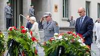 Drei Personen stehen nebeneinander vor Blumenkränzen, der Soldat in der Mitte grüßt militärisch