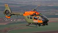Ein grüner Hubschrauber mit orangefarbener Lackierung und den Buchstaben SAR fliegt. Unter ihm liegen Felder und Dörfer.