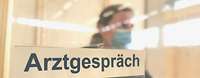 Plexiglasscheibe mit dem Schild Arztgespräch, dahinter sitzt eine Ärztin der Bundeswehr