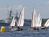 Vier kleine graue Boote segeln auf offenem Wasser, im Hintergrund ein graues Kriegsschiff.