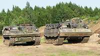 Zwei Soldaten in zwei nebeneinanderstehenden Panzern, im Hintergrund Wald 