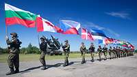 Mehrere Soldaten unterschiedlicher Nationen marschieren hintereinander und tragen verschiedene Länderflaggen