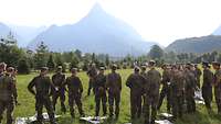 Soldaten stehen im Halbkreis auf einer Wiese vor einem anderen Soldaten, der spricht. Im Hintergrund Berge.