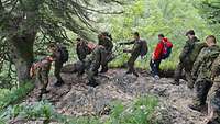 Mehrere Soldaten steigen mit einer Trage einen schmalen Gebirgsweg hinunter, unter ihnen ein Zivilist mit roter Jacke.