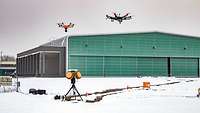 Two airborne drones in front of a hangar