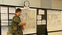 Ein Soldat steht vor einer Landkarte an einer Wand und zeigt auf etwas auf der Karte.