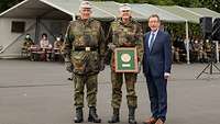 Zwei Soldaten und ein Zivilist stehen nebeneinander. Der mittlere Soldat hält einen umrahmten Ehrenpreis in der Kamera.