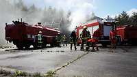 Mehrere Feuerwehrleute und rote Löschfahrzeuge stehen vor einem Flugzeughangar, aus dem Rauch tritt.