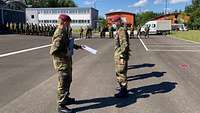 Zwei Soldaten stehen im Vordergrund. Einer übergibt eine Urkunde. 