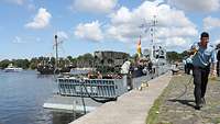 Ein graues Boot liegt an an einer Pier. Im Vordergrund ist ein Marinesoldat mit einer Leine zu sehen.