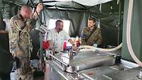 Drei Soldaten stehen in der Feldküche und unterhalten sich; einer von ihnen trägt eine Kochjacke