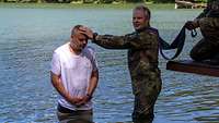 Ein Mann in weißem Shirt steht in einem See. Ein Mann in Uniform steht daneben und hält eine Hand an seinen Kopf.