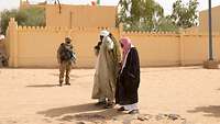 Zwei Bewohner mit traditioneller Tuaregbekleidung laufen auf einer Straße an einem Soldaten vorbei