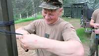 Ein deutscher Soldat knotet mit seinen Händen ein Kabel an einen Mast unter einem grünen Tarnnetz.