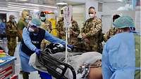 Zwei Sanitäter schieben eine Liege mit einem Verwundeten an mehreren Soldaten vorbei