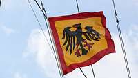 Eine gelbe Fahne mit rotem Rand und einem mittig abgebilden Adler weht im Wind.