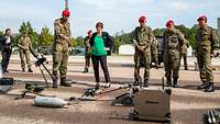 Eine Gruppe von Soldaten zeigt einer Zivilistin technisches Material, das auf einem grauen Platz im Freien liegt.