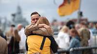 A sailor in khaki-coloured work uniform hugs a woman in a yellow and black jacket.