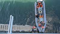 Zivilsten in Schwimmwesten steigen von einem grauen Gummiboot auf eine Leiter eines Schiffes über.