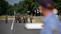 Mehrere Soldaten in komplettem Kampfanzug tragen einen verletzten Kameraden auf einem Tragetuch in Richtung Kasernentor.