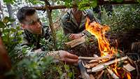 Zwei Soldaten liegen auf dem Boden, einer legt Holz in ein kleines Lagerfeuer vor ihnen.