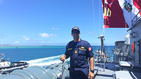 The naval officer from Peru standing on board a ship; to the left the sea, behind him the Peruvian flag is flying