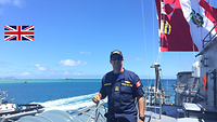 Der Marineoffizier aus Peru steht an Bord eines Schiffes. Daneben ist das Meer zu sehen, hinter ihm weht die peruanische Flagge