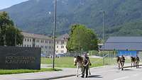 Mehrere Soldaten gehen mit ihren Maultieren hintereinander an einer Kaserne entlang, im Hintergrund Berge.