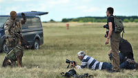 Ein Soldat steht mit seinem Diensthund auf einer Wiese neben zivilen Fotografen, die liegen und stehen.