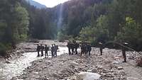 Eine Gruppe Jugendlichen laufen durch ein Flussbett, im Hintergrund Wald und Berge.