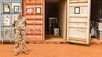 Ein Soldat mit blauem Barett und Tarnuniform steht vor drei Containern mit Material