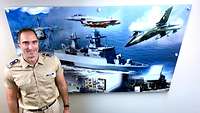 A soldier is smiling and standing in front of a wall with a poster that shows two ships and an aircraft