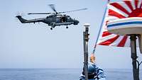 Ein Marinesoldat in hellblauer Uniform winkt mit einer rot-weißen Flagge einem Hubschrauber.