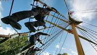 3 Personen in blauer Uniform klettern die Strickleitern an einem Mast empor.