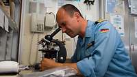 Ein Mann in blauer Uniform sitzt an einem Tisch und schaut durch ein Mikroskop.