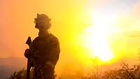 Ein Soldat in Gefechtsanzug steht bei Sonnenuntergang auf einer Wiese.