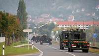 Auf einer Landstraße fahren drei Kampfpanzer hintereinander, zwei Tieflader folgen, im Hintergrund Häuser am Hang.