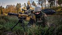 Ein EGB-Soldat liegt mit Maschienengewehr im Anschlag im Gelände, im Hintergrund ein Hubschrauber am Boden