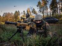 Ein EGB-Soldat liegt mit Maschienengewehr im Anschlag im Gelände, im Hintergrund ein Hubschrauber am Boden