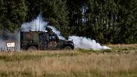 Ein geschütztes Fahrzeug steht auf einer Wiese vor einer Waldkante. Vor dem Fahrzeug befindet sich eine Rauchkartusche.