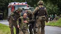 Zwei Soldaten helfen einem Übungsverletzten, der kniet. Im Hintergrund ist ein Feuerwehrfahrzeug zu sehen.