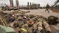 Soldaten liegen auf einem großen Platz vor Lastkraftwagen auf dem Boden und bedecken Kopf und Nacken schützend mit ihren Händen.