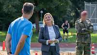 Ein Mann in blauem Sportshirt spricht mit einer Zivilistin. Sie stehen an einer Sprunggrube mit Sand.