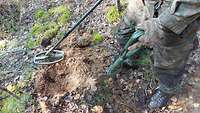Ein Soldat steht mit einem Metalldetektor in der rechten Hand und einem Klappspaten in der linken Hand im Wald.