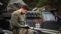 Ein Soldat sitzt mit einem Buch auf der Motorhaube eines Fahrzeugs und schreibt etwas hinein.