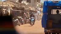 Viele Motorroller stehen am Straßenrand. Ein Mann mit dunkler Hautfarbe fährt auf einer Straße aus Sand