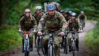 Mehrere Soldaten auf dem Fahrrad kommen einen Waldweg hinaufgefahren.