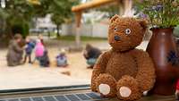 Ein Teddybär auf einem Fensterbrett, dahinter Kinder in einem Garten